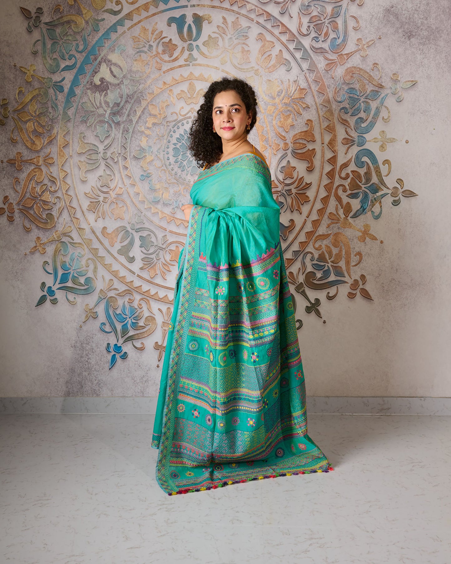 Woman wearing a turquoise colour chanderi silk cotton saree with Lambani embroidery all over the saree standing in front of a beautiful backdrop with a mandala design on it.