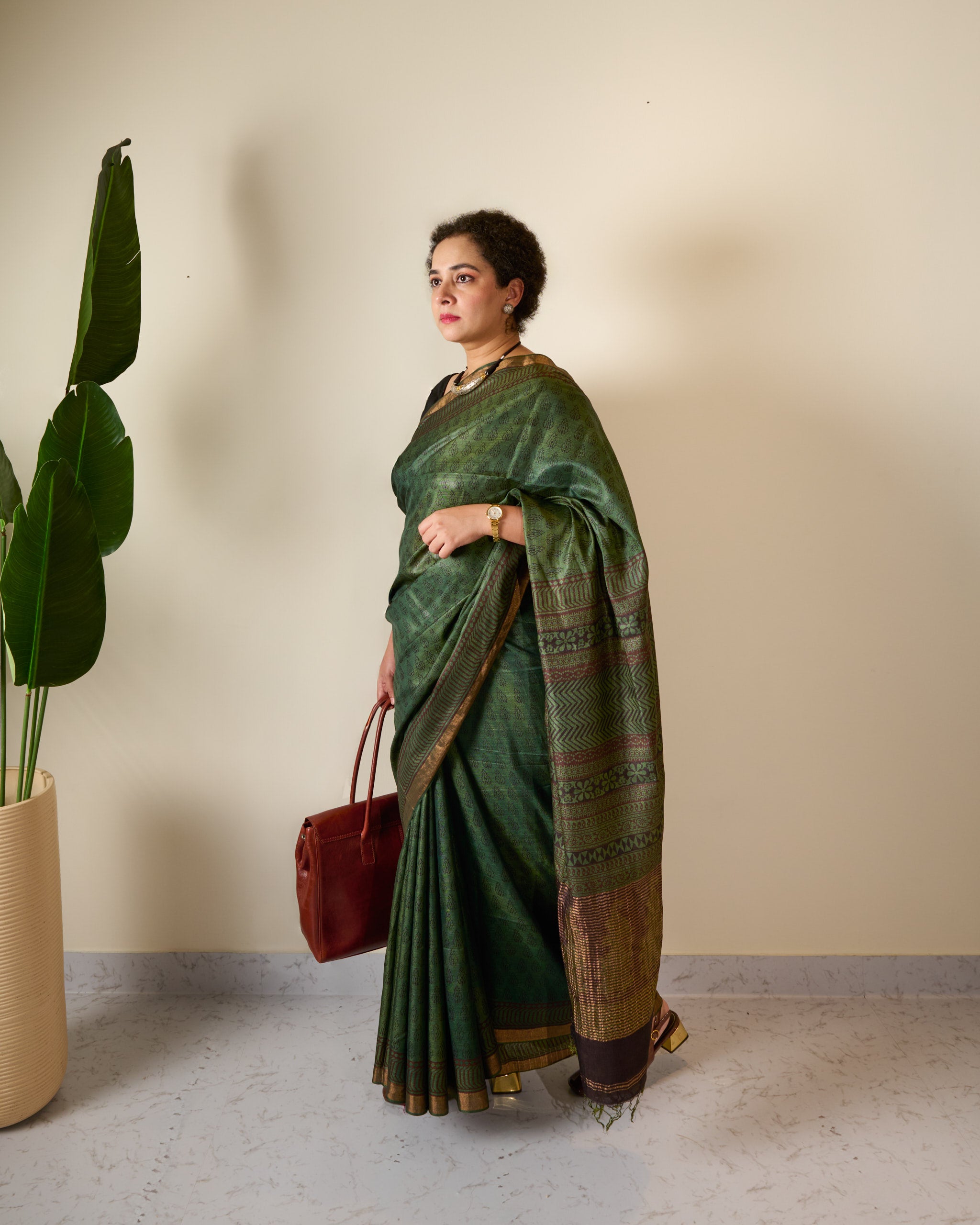 Woman wearing forest green Kosa silk saree with block prints all over the saree and the pallu standing in front of a cream with a handbag in one hand, seems to be walking towards somewhere.