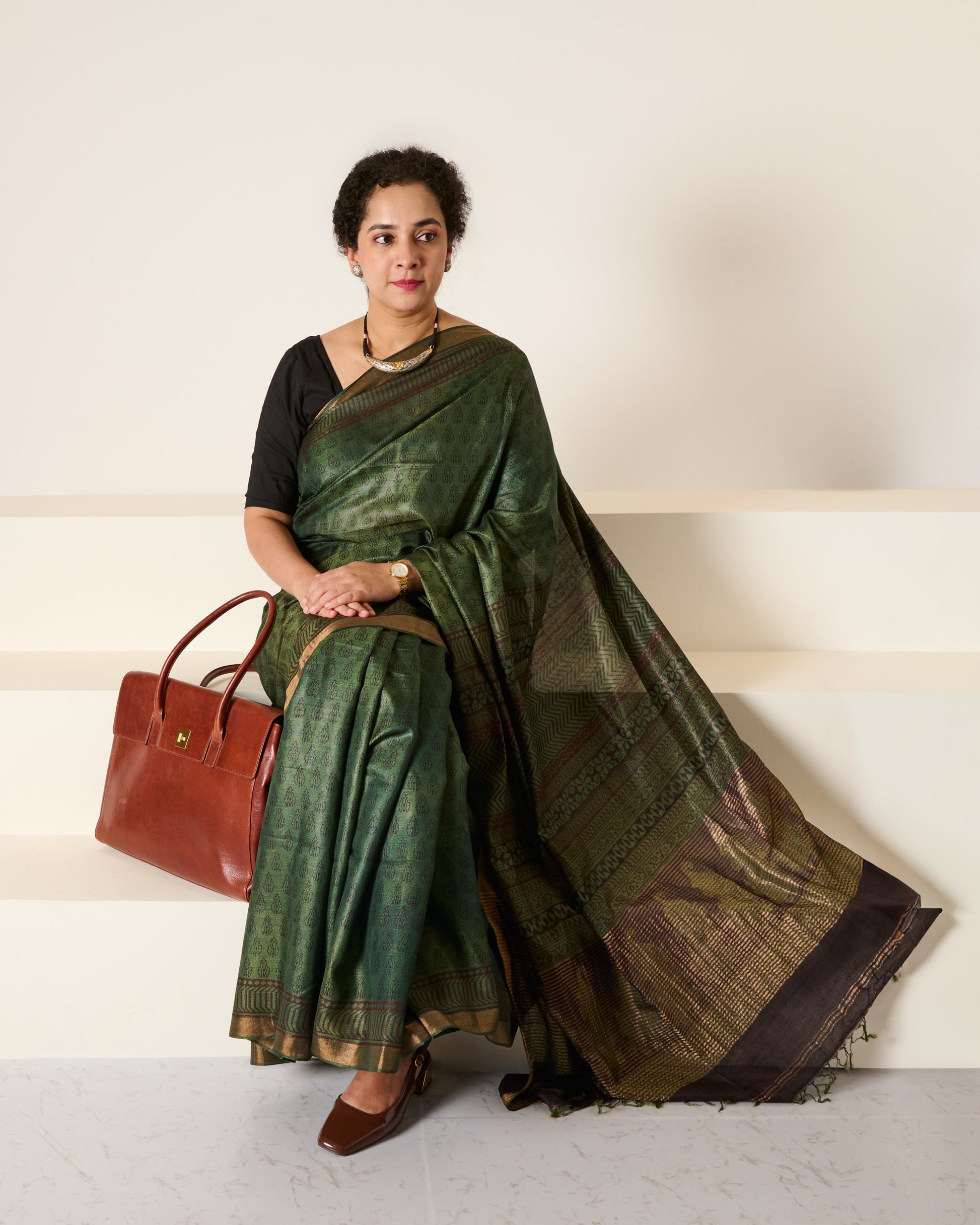 Woman wearing forest green Kosa silk saree with block prints all over the saree and the pallu standing in front of a cream wall with a handbag to her side sitting on white stairs, with her hands folded on her lap. 