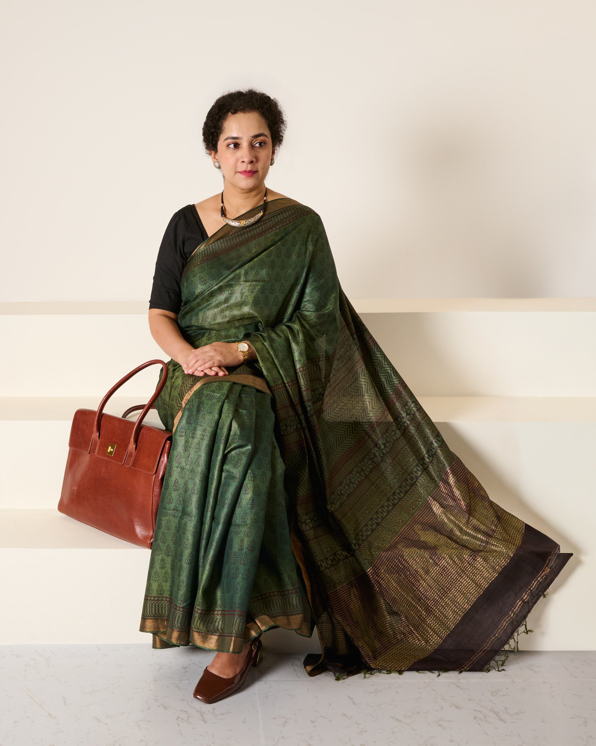 Woman wearing forest green Kosa silk saree with block prints all over the saree and the pallu standing in front of a cream wall with a handbag to her side sitting on white stairs, with her hands folded on her lap. 