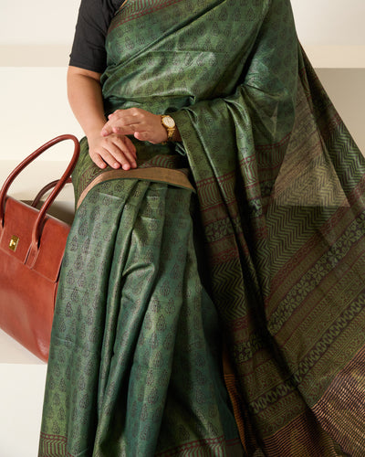 Woman wearing forest green Kosa silk saree with block prints all over the saree and the pallu sitting on white stairs.