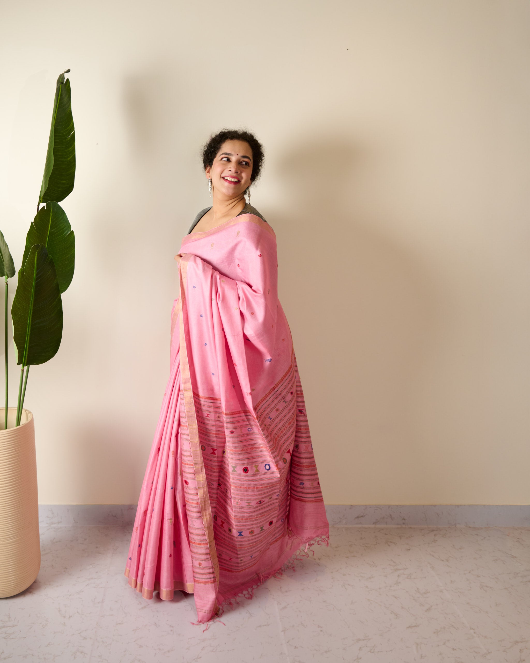 Woman wearing a pink kosa silk saree standing next to a plant against a plain wall.
