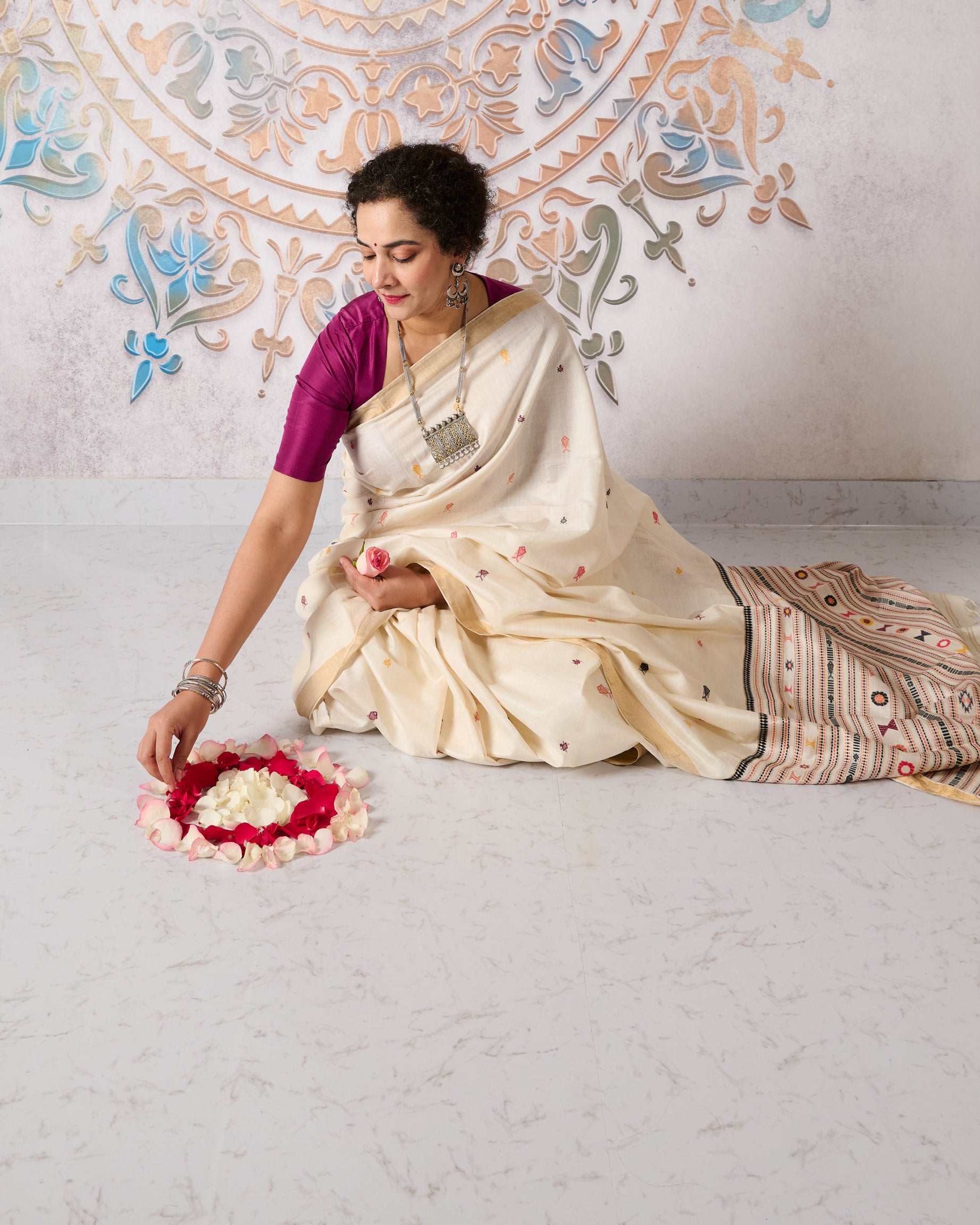 Woman in a traditional kosa silk saree in white with multi colour motifs performing a ritual with a floral arrangement on a light background