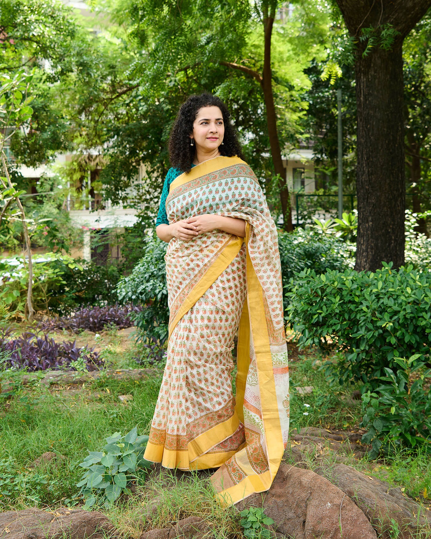 Woman wearing a block printed kota silk saree in white base with orange flowers standing amidst shrubs and flowers.