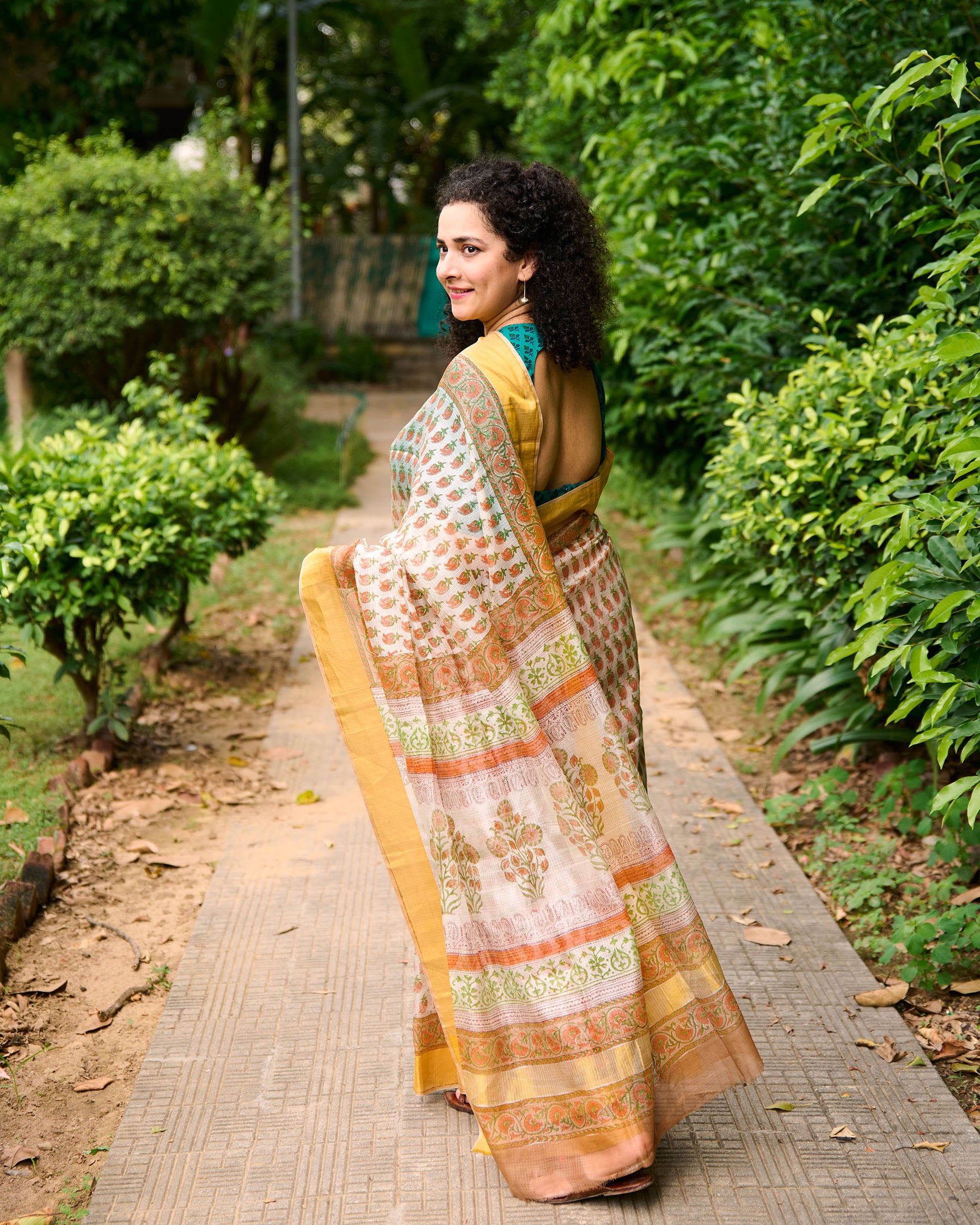 Woman wearing a block printed kota silk saree in white base with orange flowers standing walking with the pallu of the saree flying behind her.