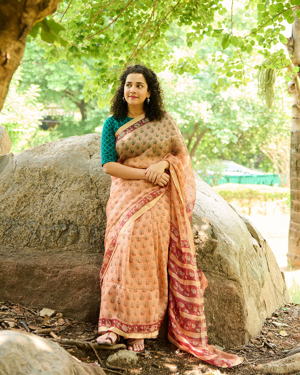 Woman wearing a peach colour kota silk saree with floral block print over a teal green blouse sitting on a rock in a forest.