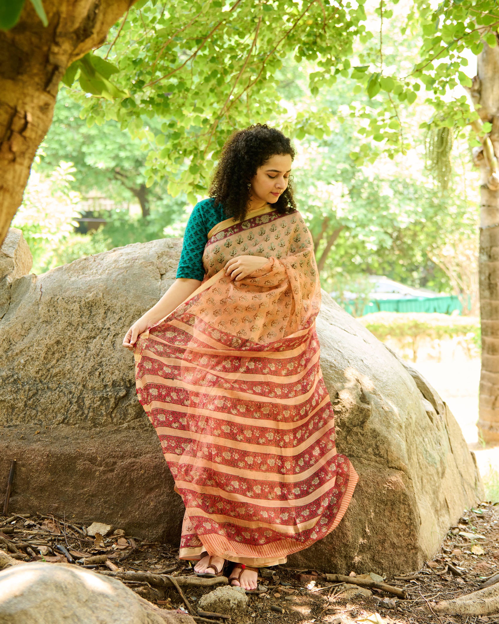 Woman wearing a peach colour kota silk saree with floral block print over a teal green blouse sitting on a rock in a forest.