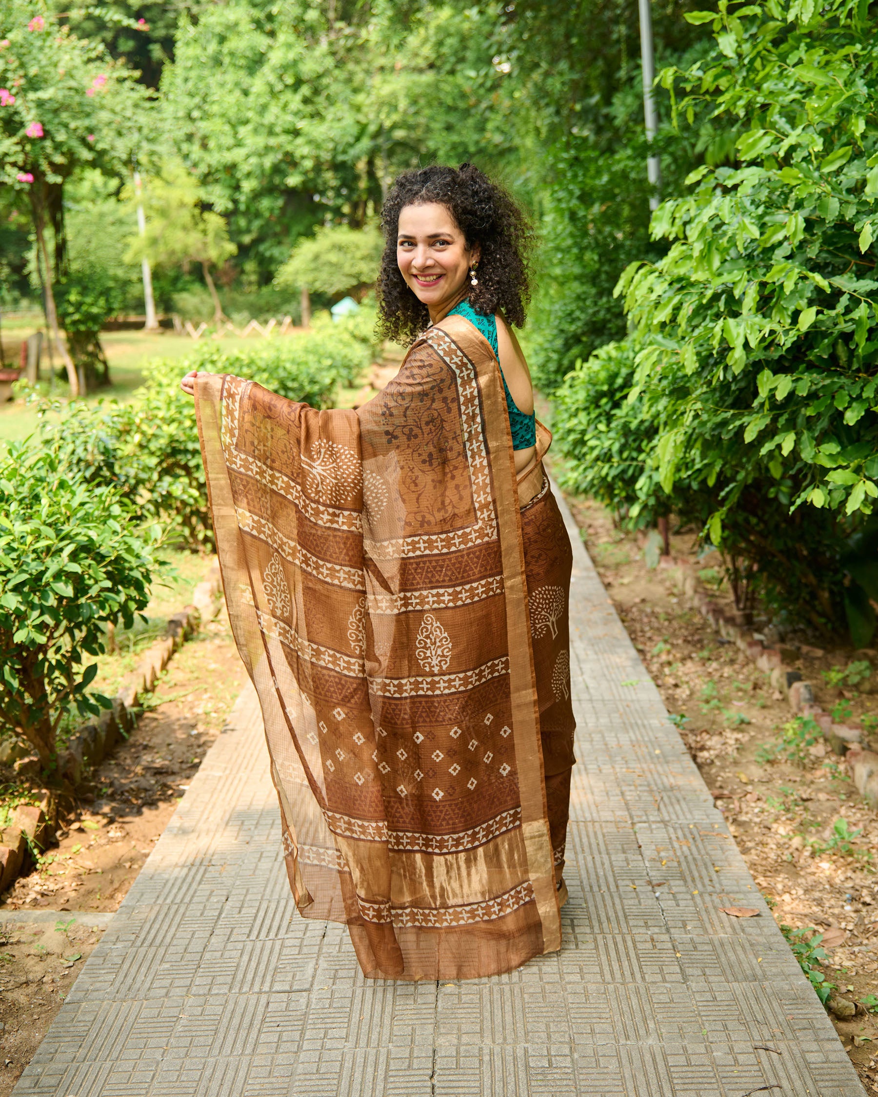 Woman wearing a brown dabu block print kota silk saree playfully strolling down a pavement in a park. 