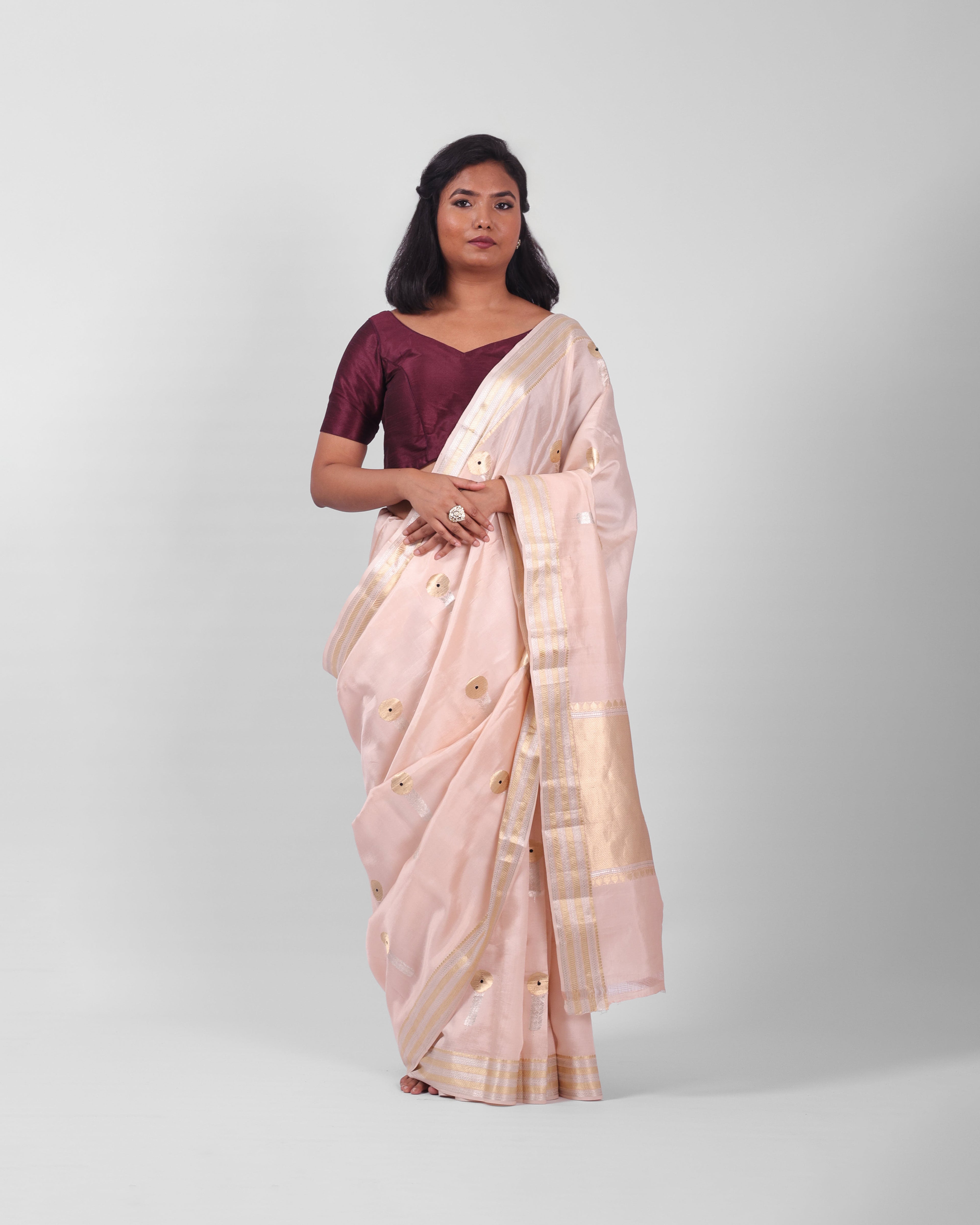 A woman modeling a blush pink banarasi kadwa saree with golden and silver zari borders and a zari minakari marigold motifs all over the saree, and an ornate pallu. 

