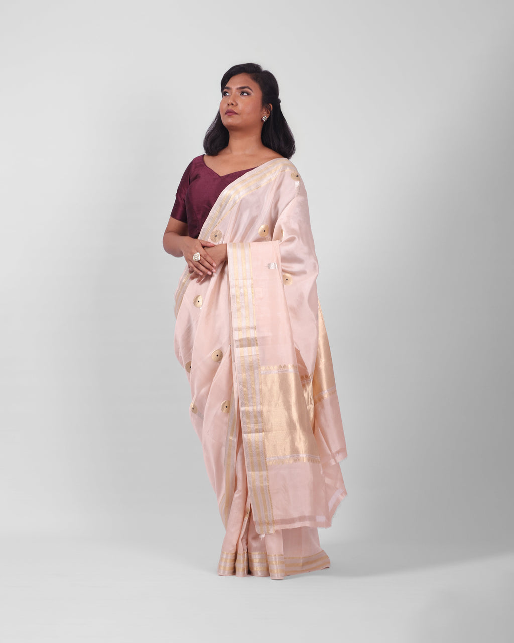 A woman modeling a blush pink banarasi kadwa saree with golden and silver zari borders and a zari minakari marigold motifs all over the saree, and an ornate pallu. 
