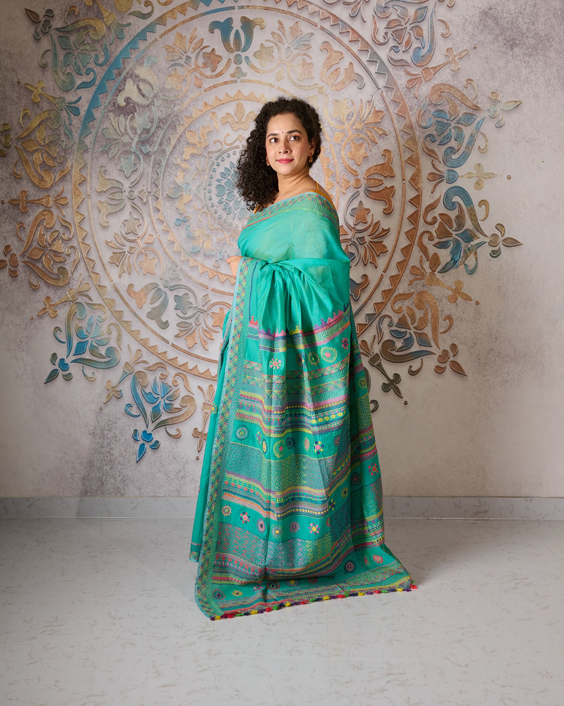 Woman wearing a turquoise colour chanderi silk cotton saree with Lambani embroidery all over the saree standing in front of a beautiful backdrop with a mandala design on it. 