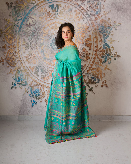 Woman wearing a turquoise colour chanderi silk cotton saree with Lambani embroidery all over the saree standing in front of a beautiful backdrop with a mandala design on it. 