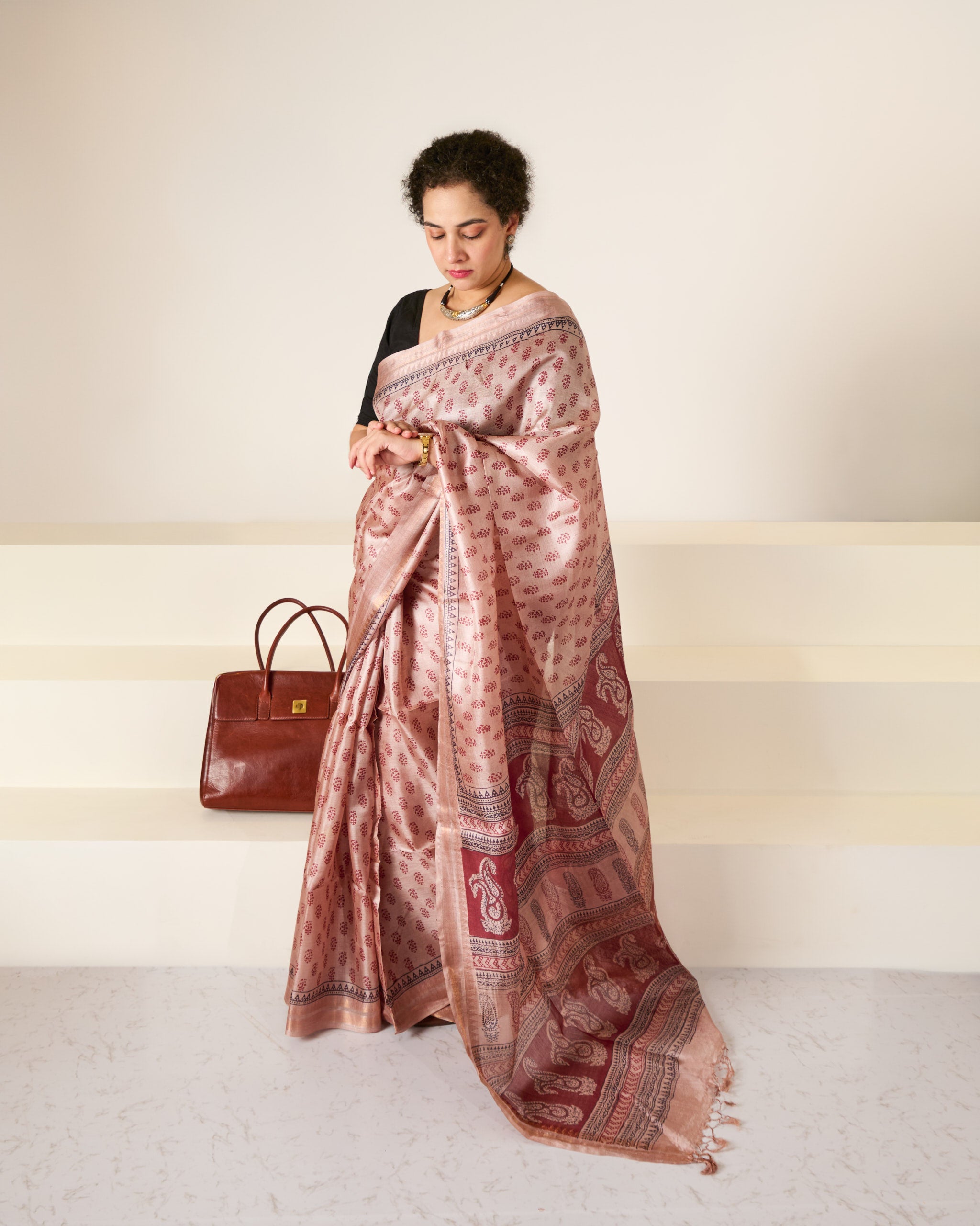 Woman wearing a dusky rose blockprinted Kosa Silk saree in front of a white backdrop and white stairs with her handbag gently seated behind her and she is looking at her watch.