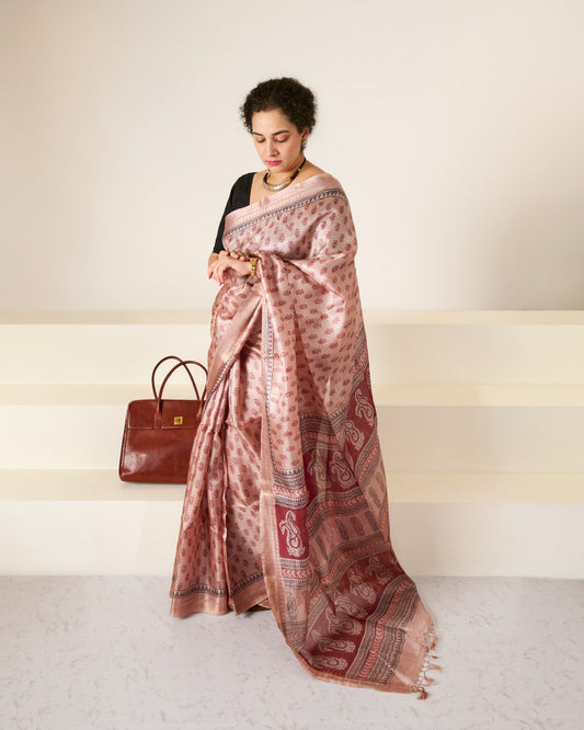 Woman wearing a dusky rose blockprinted Kosa Silk saree in front of a white backdrop and white stairs with her handbag gently seated behind her and she is looking at her watch.