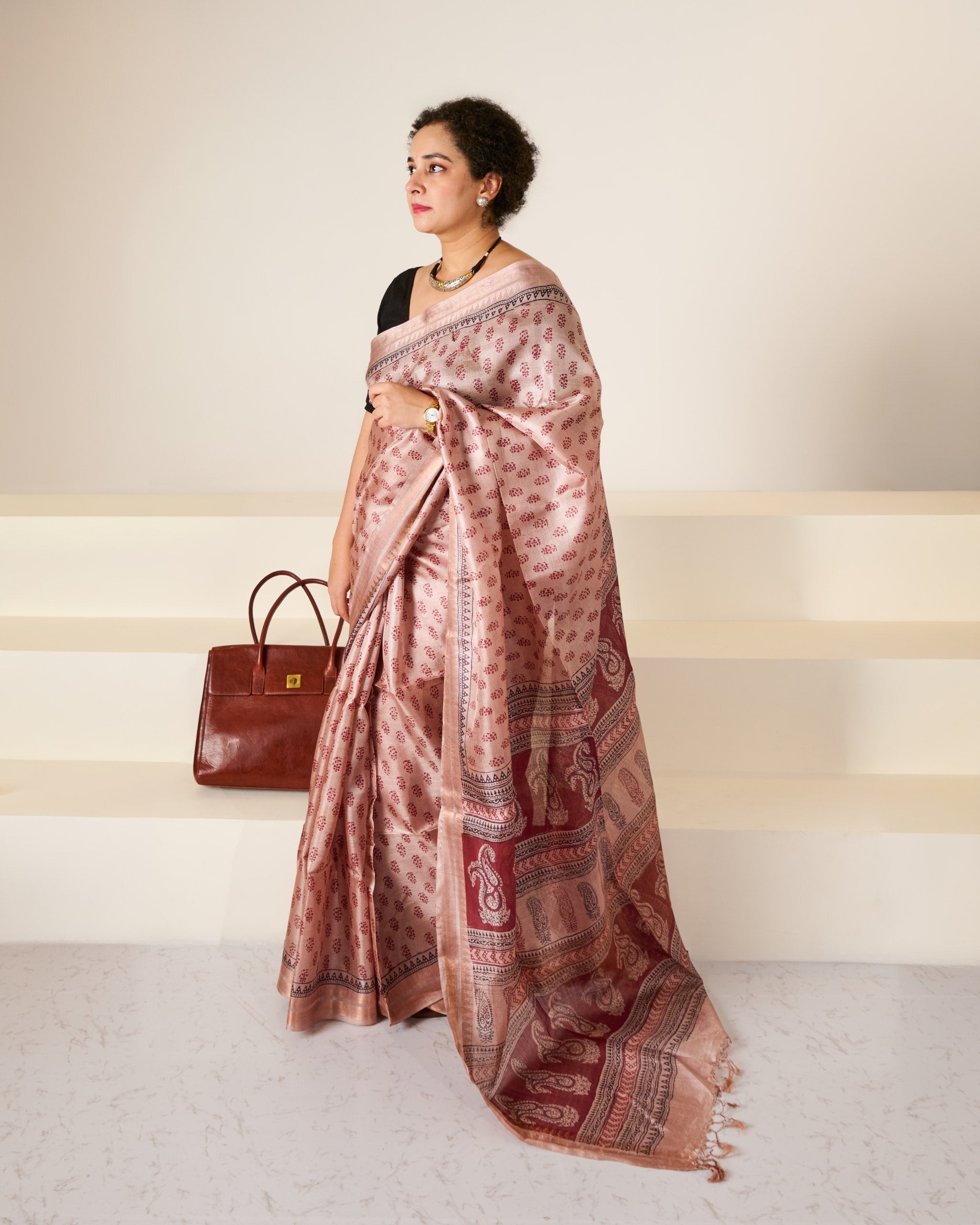 Woman wearing a dusky rose blockprinted Kosa Silk saree in front of a white backdrop and white stairs with her handbag gently seated behind her.