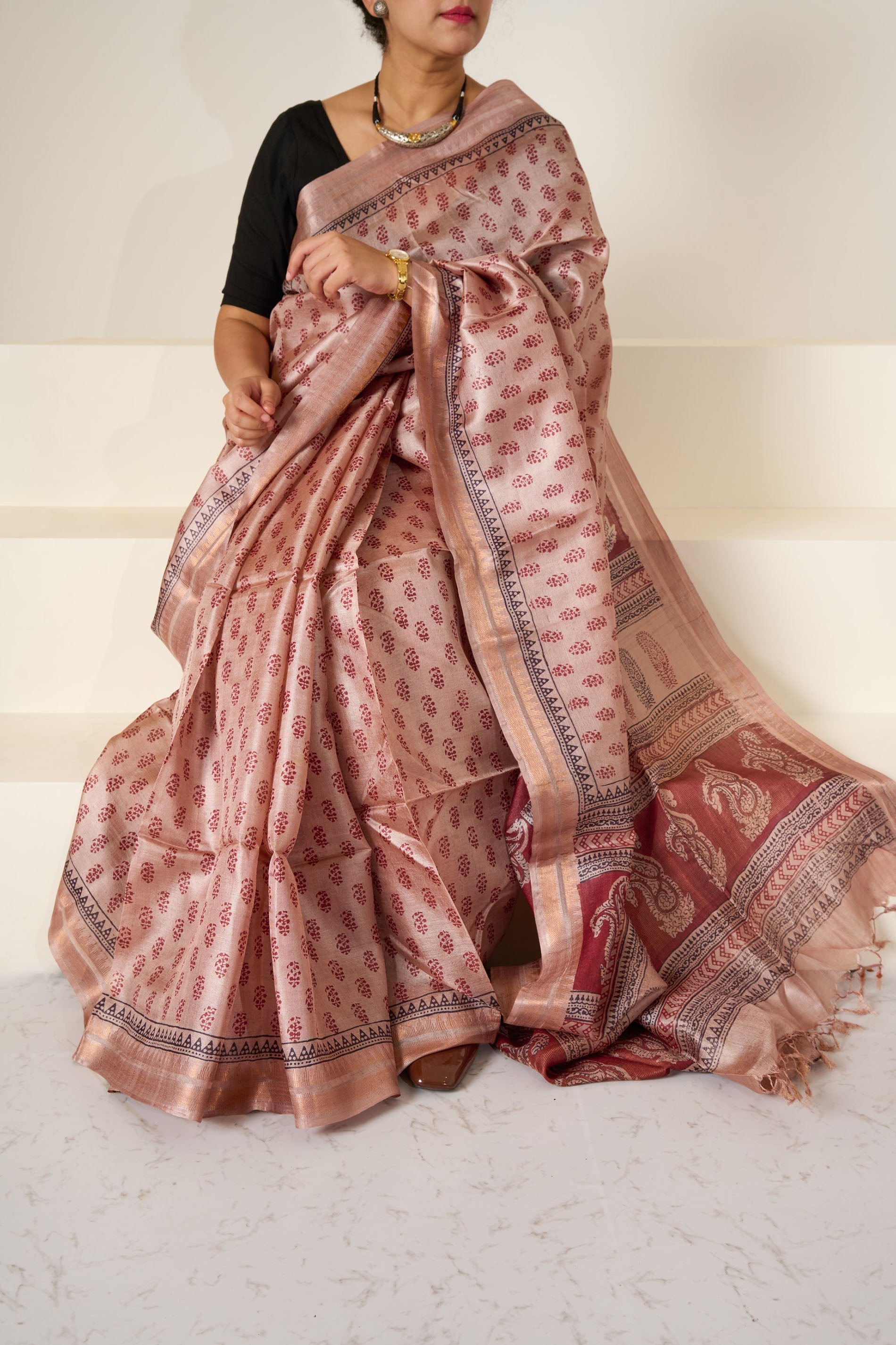 Woman wearing a dusky rose blockprinted Kosa Silk saree in front of a white backdrop sitting on white stairs.