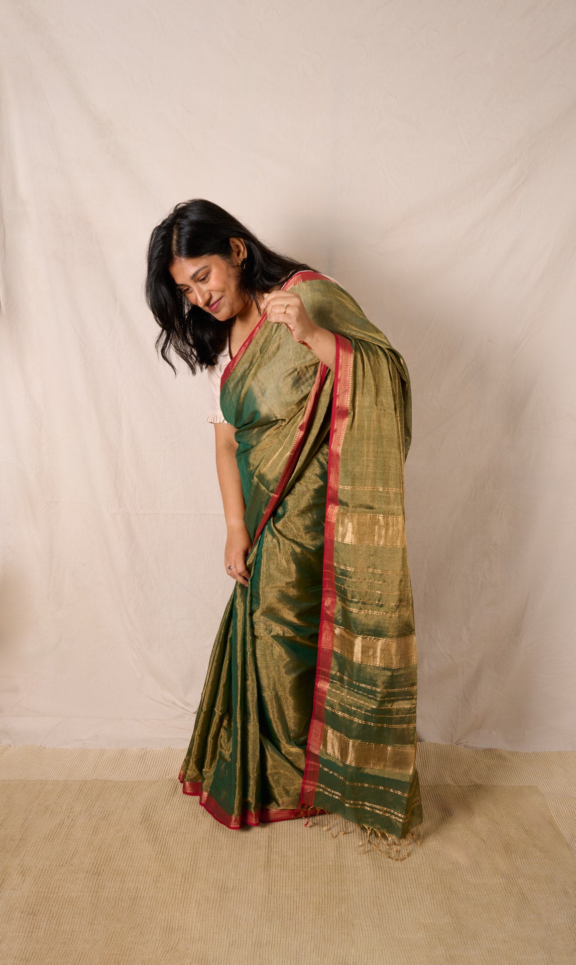 Woman wearing a maheshwari tissue saree in dual tone of dark green and gold standing in front of a beige background adjusting her pleats.