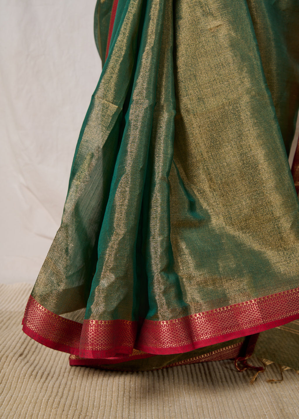 Neat pleats of a maheshwari tissue saree in dual tone of dark green and gold standing in front of a beige background.