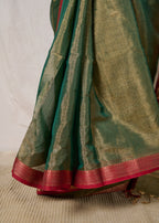 Neat pleats of a maheshwari tissue saree in dual tone of dark green and gold standing in front of a beige background.