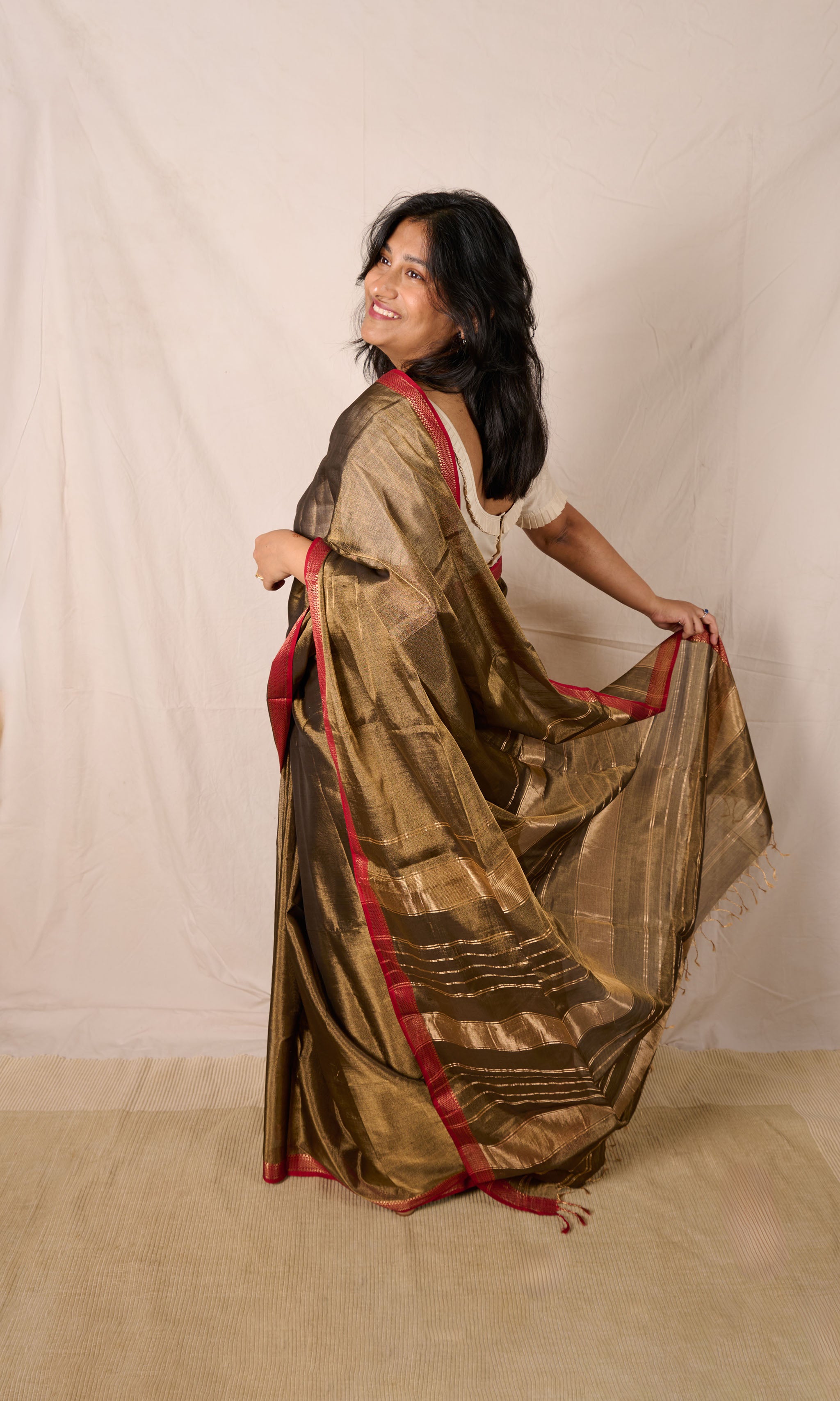 Women wearing a maheshwari tissue saree in dual tone of black and gold standing in front a beige backdrop. 