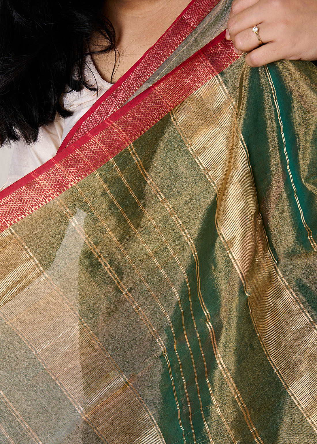 Woman wearing a maheshwari tissue saree in dual tone of dark green and gold standing in front of a beige background showcasing the ornate contrast red border.