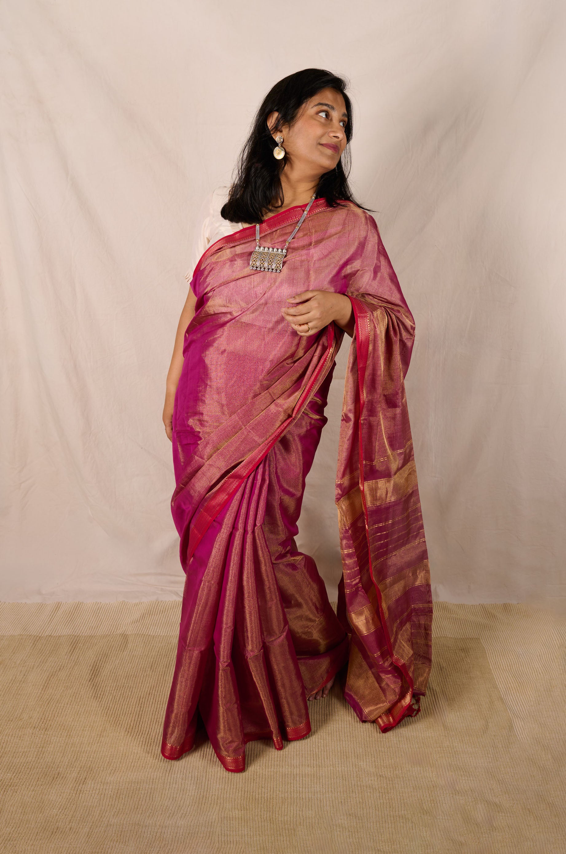 Women wearing a maheshwari tissue saree in dual tone of pink and gold standing in front a beige backdrop. 