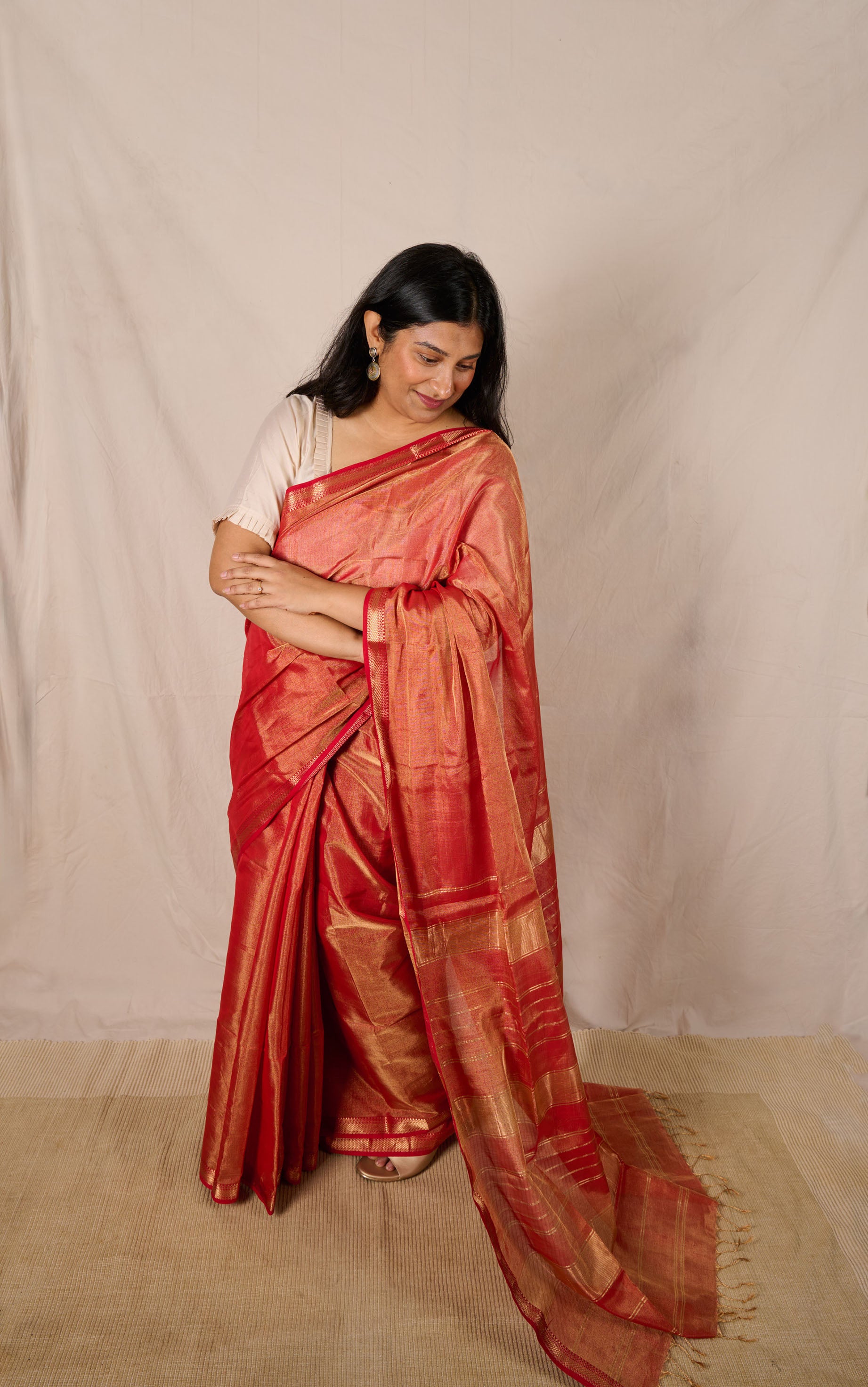 Woman wearing a maheshwari tissue saree in dual tone of red and gold standing in front a beige backdrop. 