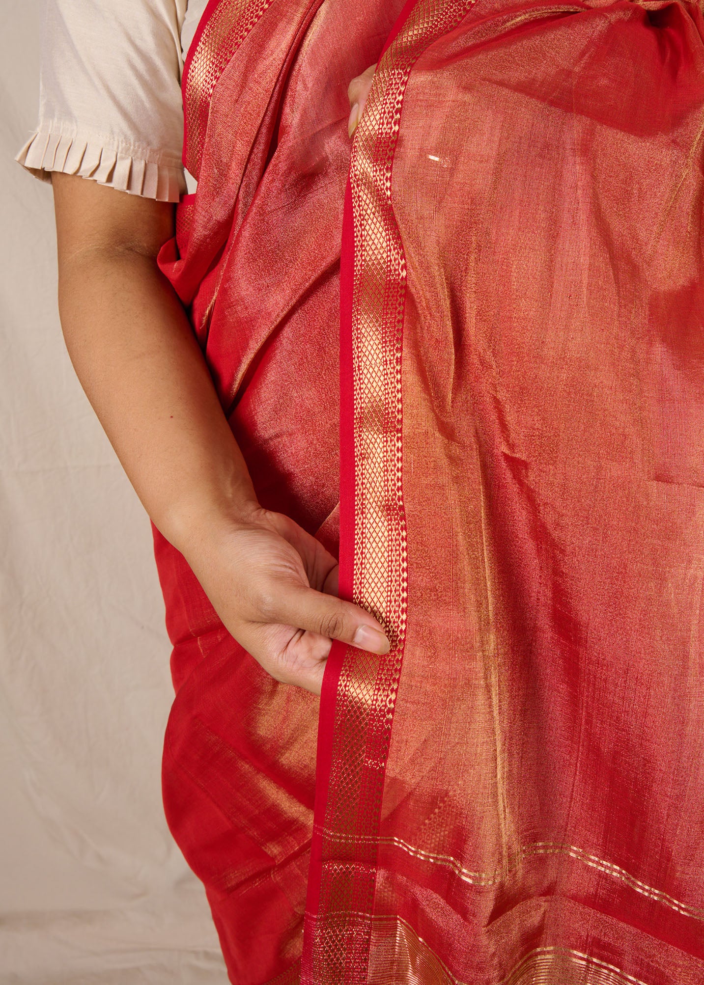 Woman wearing a maheshwari tissue saree in dual tone of red and gold standing in front a beige backdrop showcasing the ornate border of the saree.