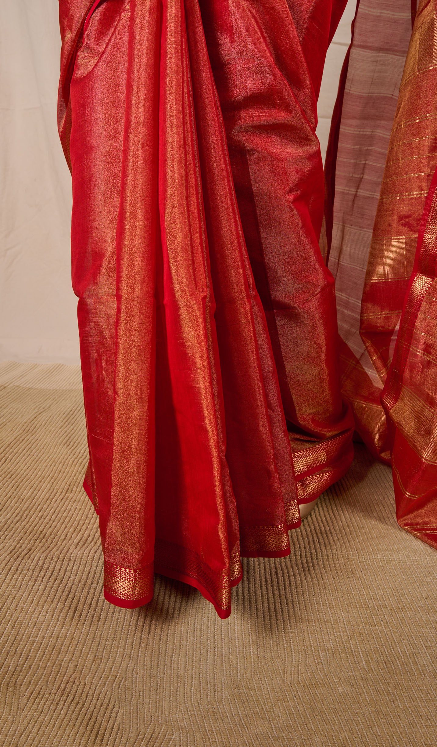 Close up of the saree pleats worn by a woman wearing a maheshwari tissue saree in dual tone of red and gold standing in front a beige backdrop 