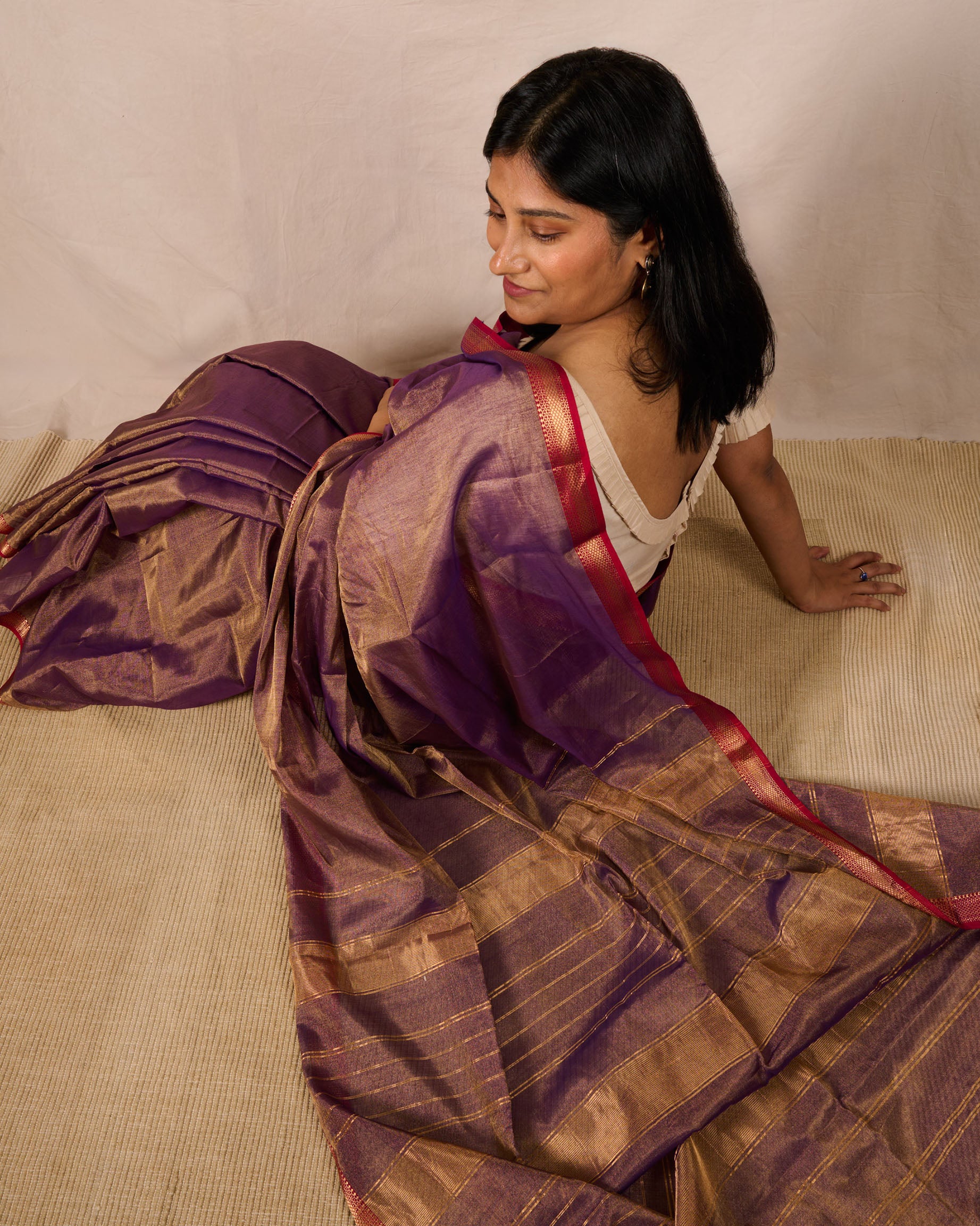 Woman wearing a dual tone Maheshwari Tissue saree in violet and gold sitting on a carpet looking over her shoulder while 