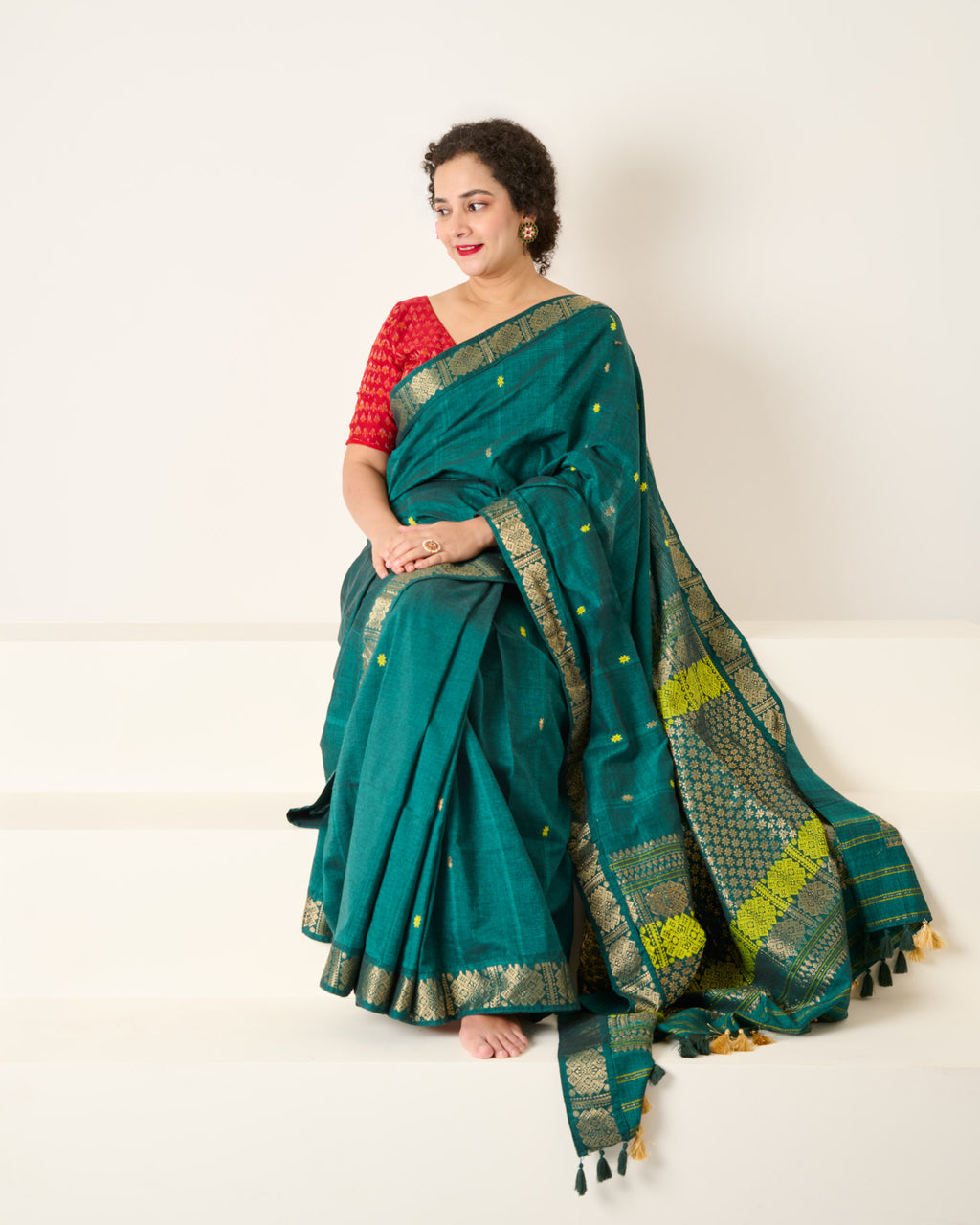 Woman is sitting on white stairs wearing a Assam cotton green saree with an ornate zari border paired with a red blouse 