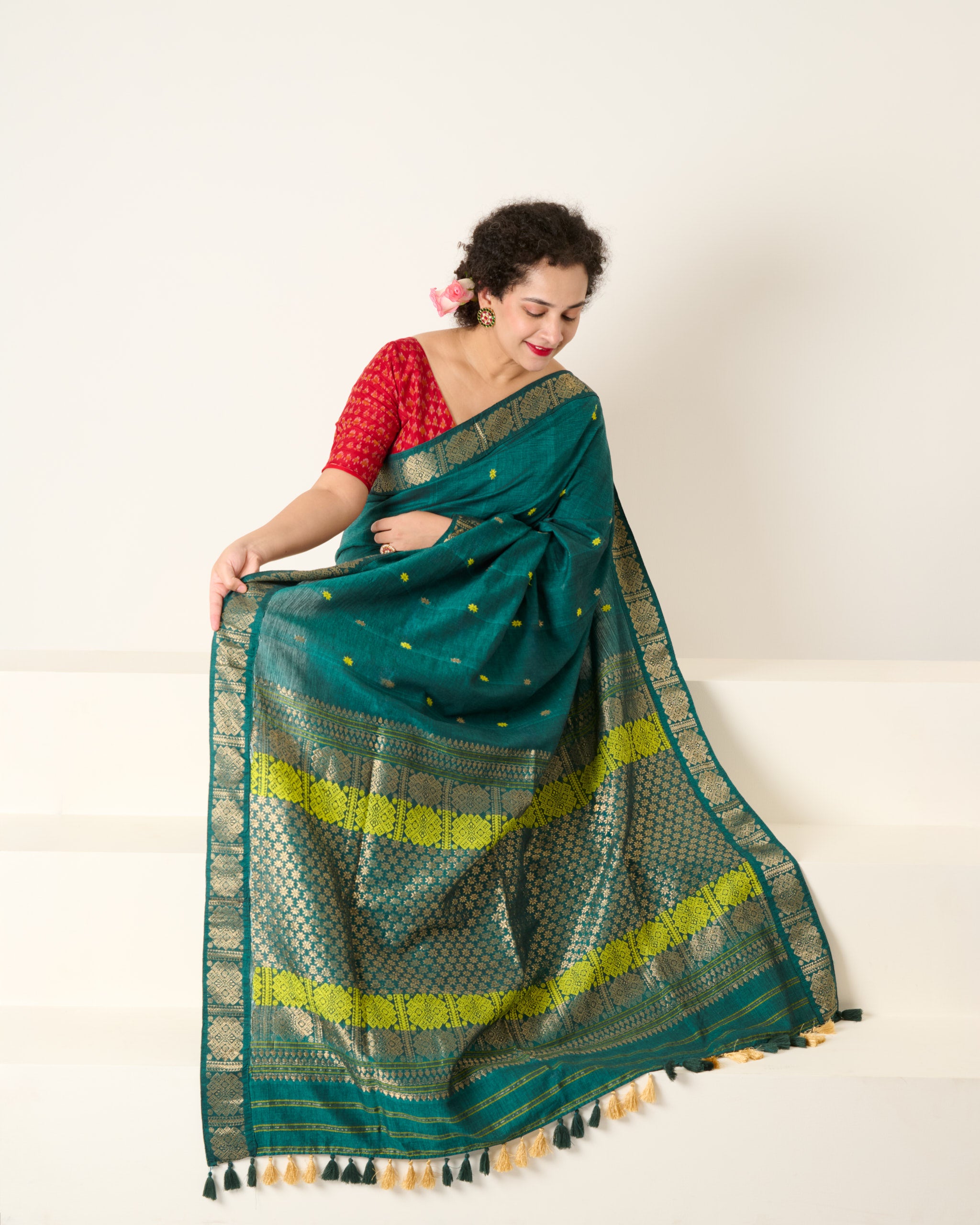 Woman is sitting on white stairs wearing a Assam cotton green saree with an ornate zari border paired with a red blouse 