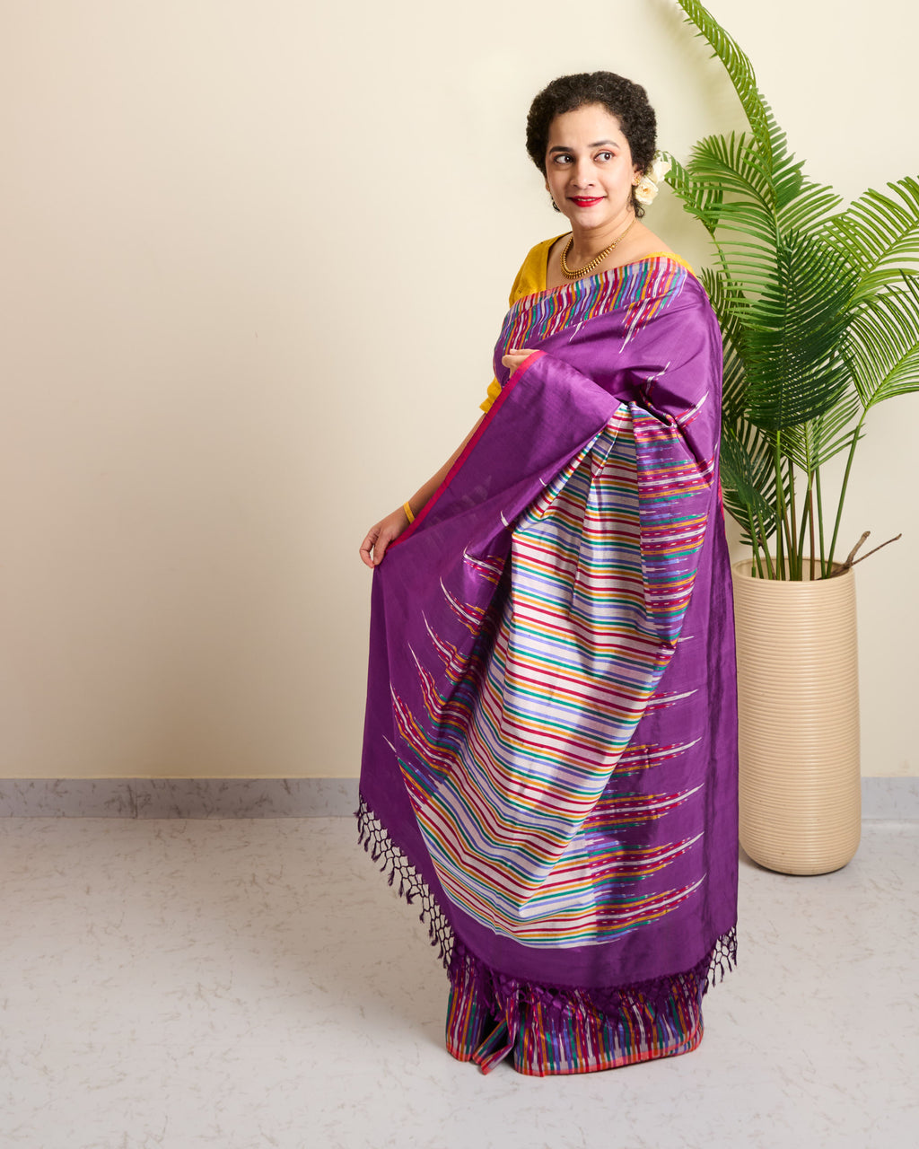 Woman wearing a purple pochampalli ikat eri silk saree with colorful patterns, standing indoors next to a plant.

