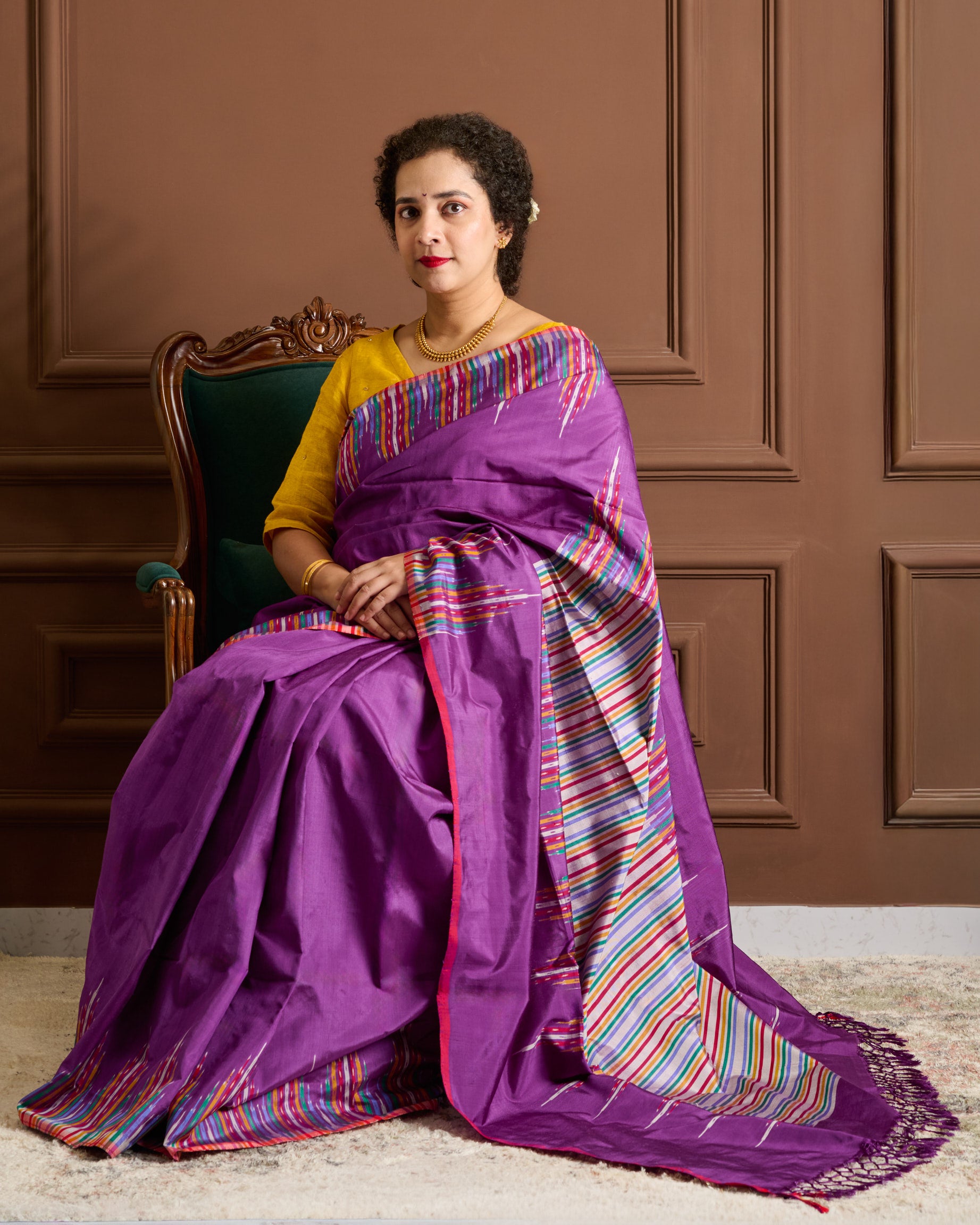 Woman wearing a purple pochampalli ikat saree with colorful patterns sitting on a chair.

