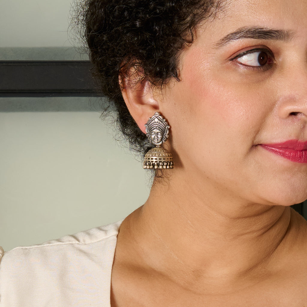Closeup of a woman wearing silver jhumka with top half engarved with Ma Durga's likeness followed by an engraved jhumka with tiny ghungroo.