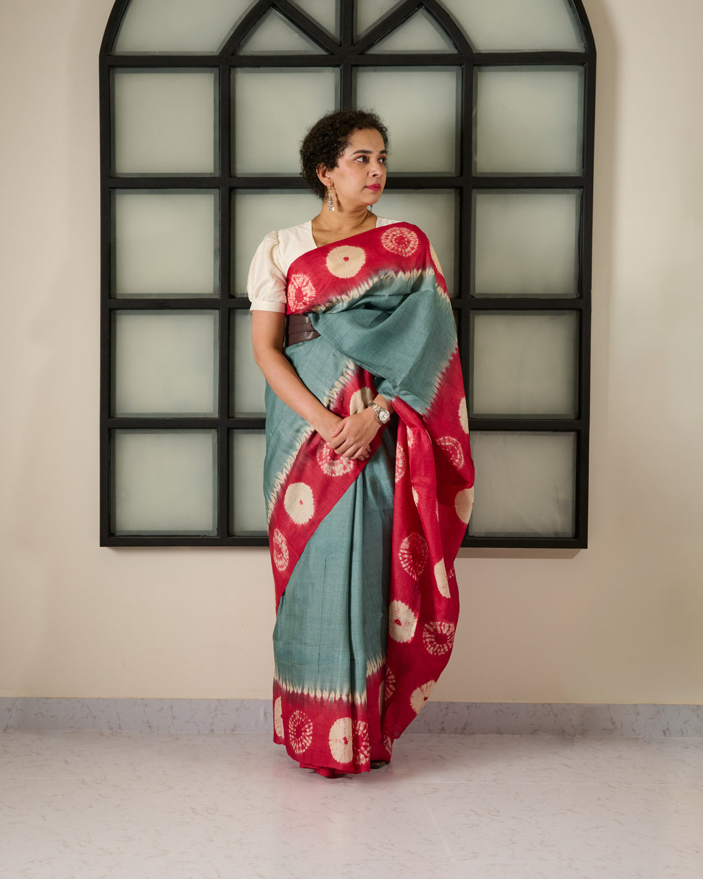 Woman wearing a tussar saree with grey body and red shibori on the border and pallu accessorised with a belt at her waist and a watch on her wrist standing against a window facade.