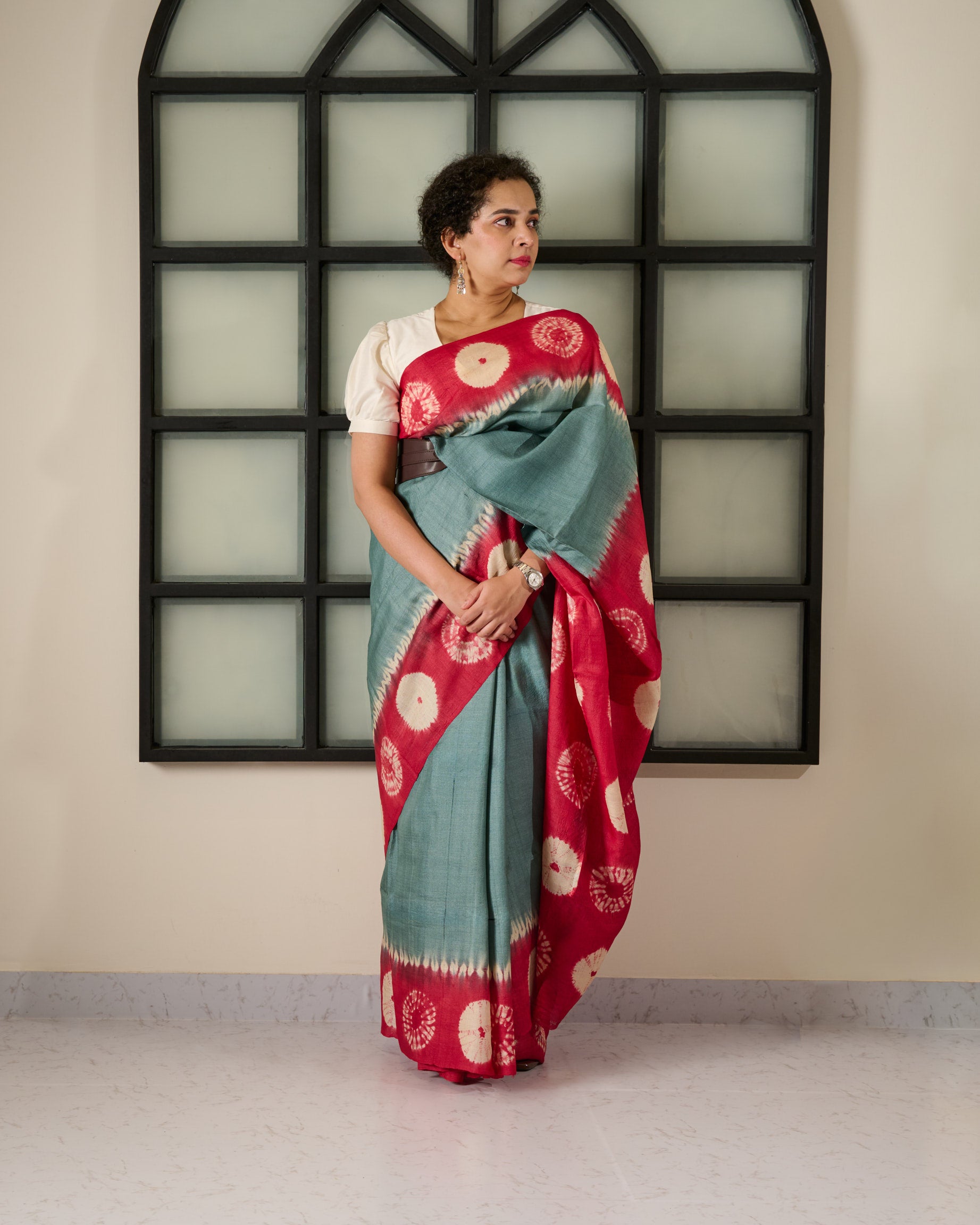 Woman wearing a tussar saree with grey body and red shibori on the border and pallu accessorised with a belt at her waist and a watch on her wrist standing against a window facade.