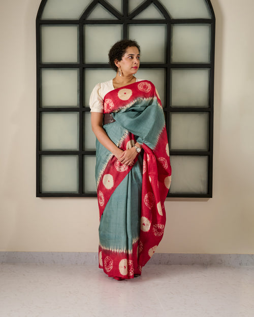 Woman wearing a tussar saree with grey body and red shibori on the border and pallu accessorised with a belt at her waist and a watch on her wrist standing against a window facade.