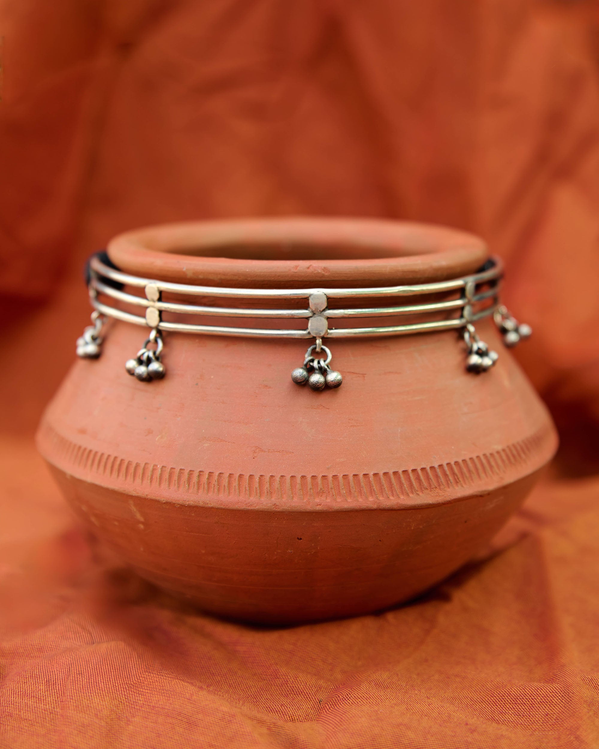 Silver hasli necklace with small hanging ghungroo charms on a terracotta pot against an orange fabric background.