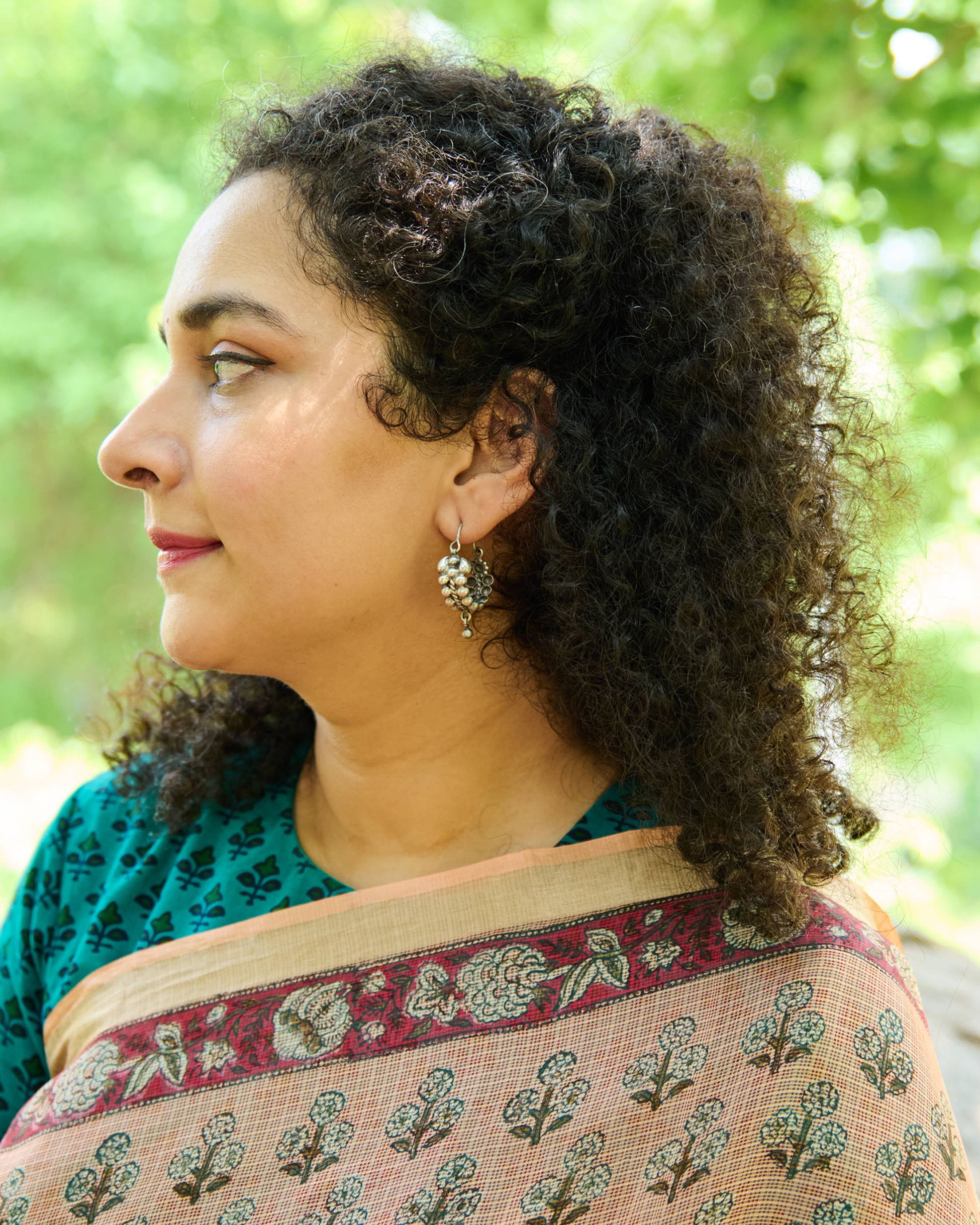 Woman wearing a block printed pink saree in a green outdoor space with her left profile towards the camera showing a silver earring in her ears.