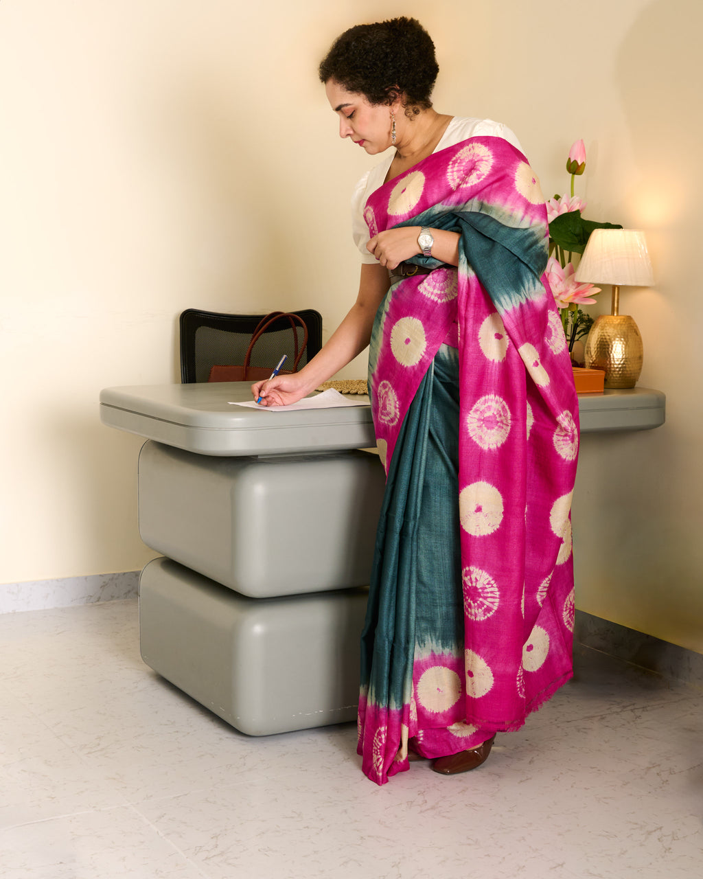 Woman wearing tussar saree with a grey body and pink shibori patterns on the border and pallu standing at her work desk