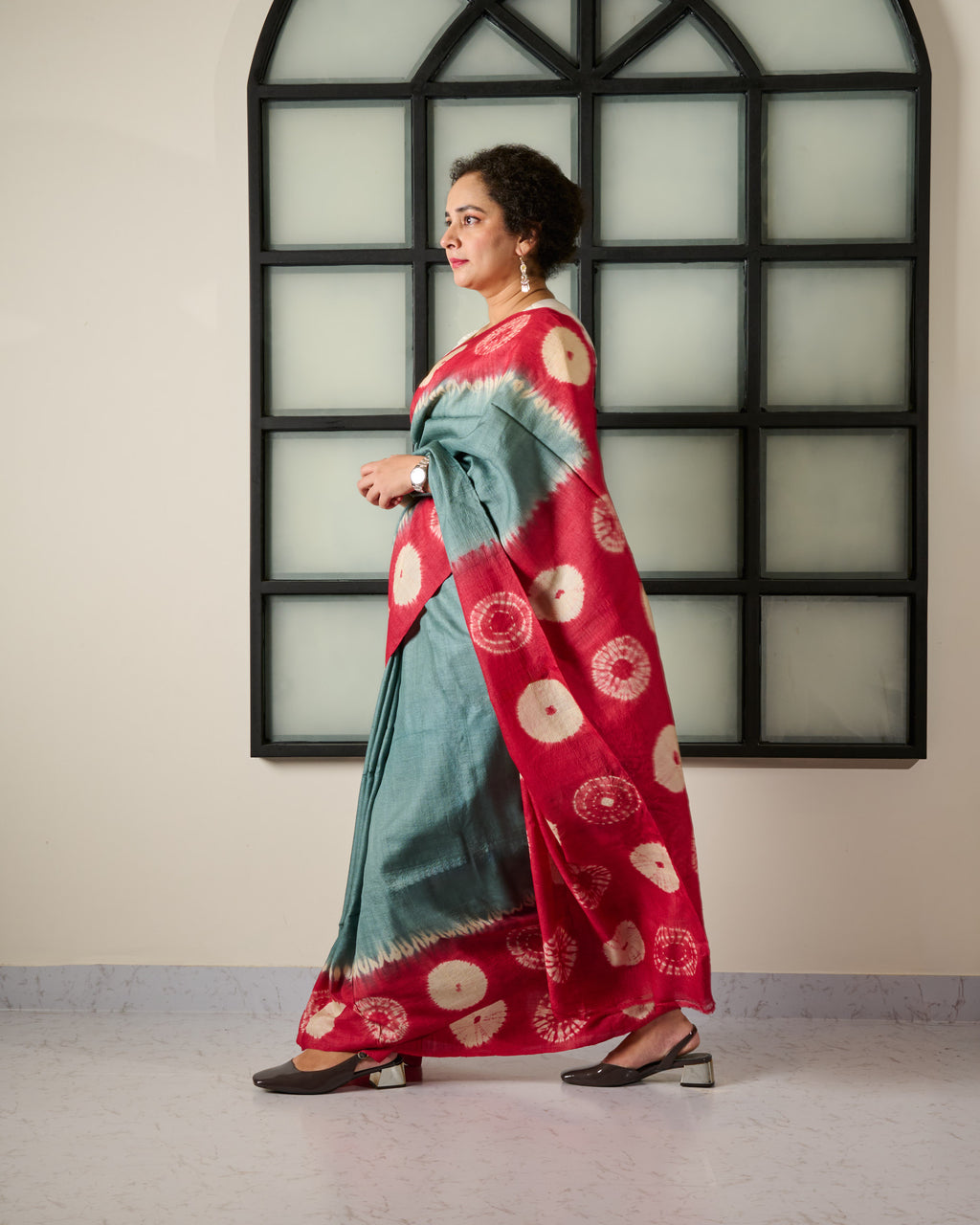 Woman wearing a tussar saree with grey body and red shibori on the border and pallu accessorised with a belt at her waist and a watch on her wrist standing against a window facade.