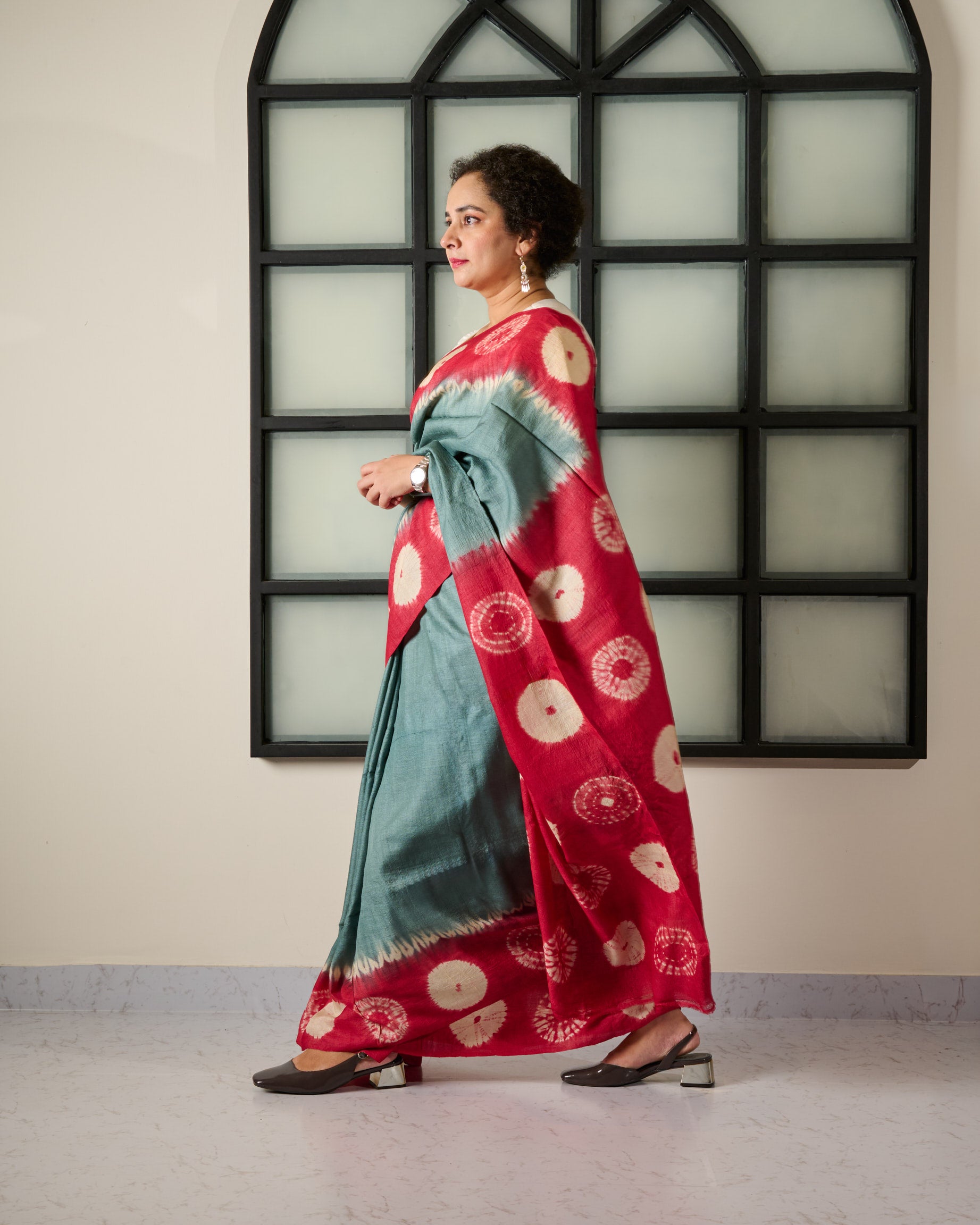 Woman wearing a tussar saree with grey body and red shibori on the border and pallu accessorised with a belt at her waist and a watch on her wrist standing against a window facade.