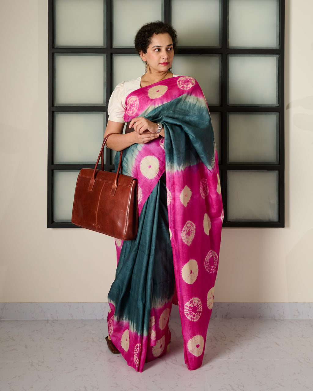 Woman wearing tussar saree with a grey body and pink shibori patterns on the border and pallu with a brown work bag in her right arm.