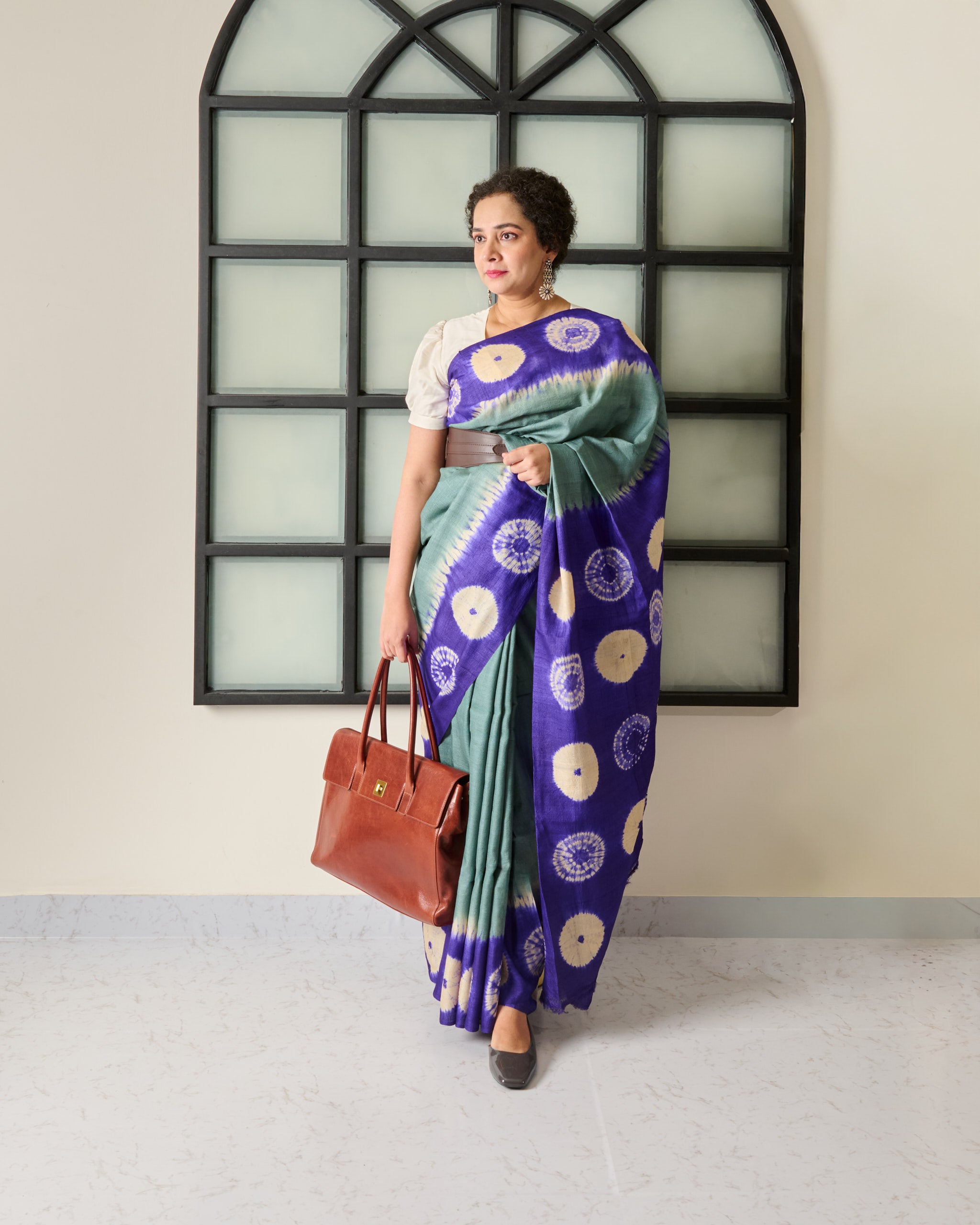 Woman holding a brown handbag wearing a tussar saree with a grey body and a purple shibori pattern on the border and pallu.