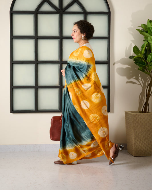 Woman wearing in a tussar saree with grey body and yellow shibori pattern on the border and pallu walking with a bag in hand.